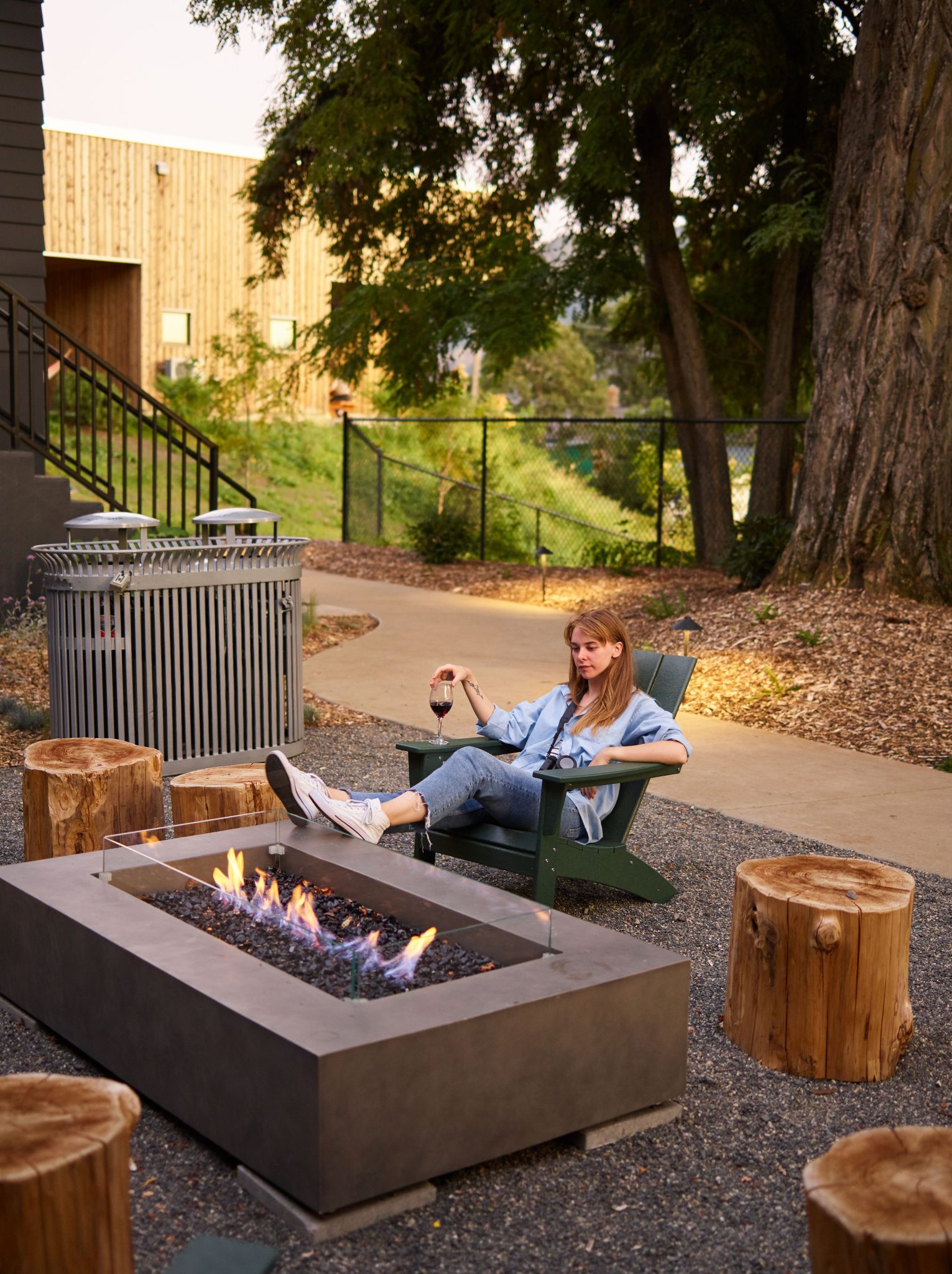 Full body lady in casual outfit chilling on garden chair near creative cozy fireplace and decorative tree stubs in summer garden
