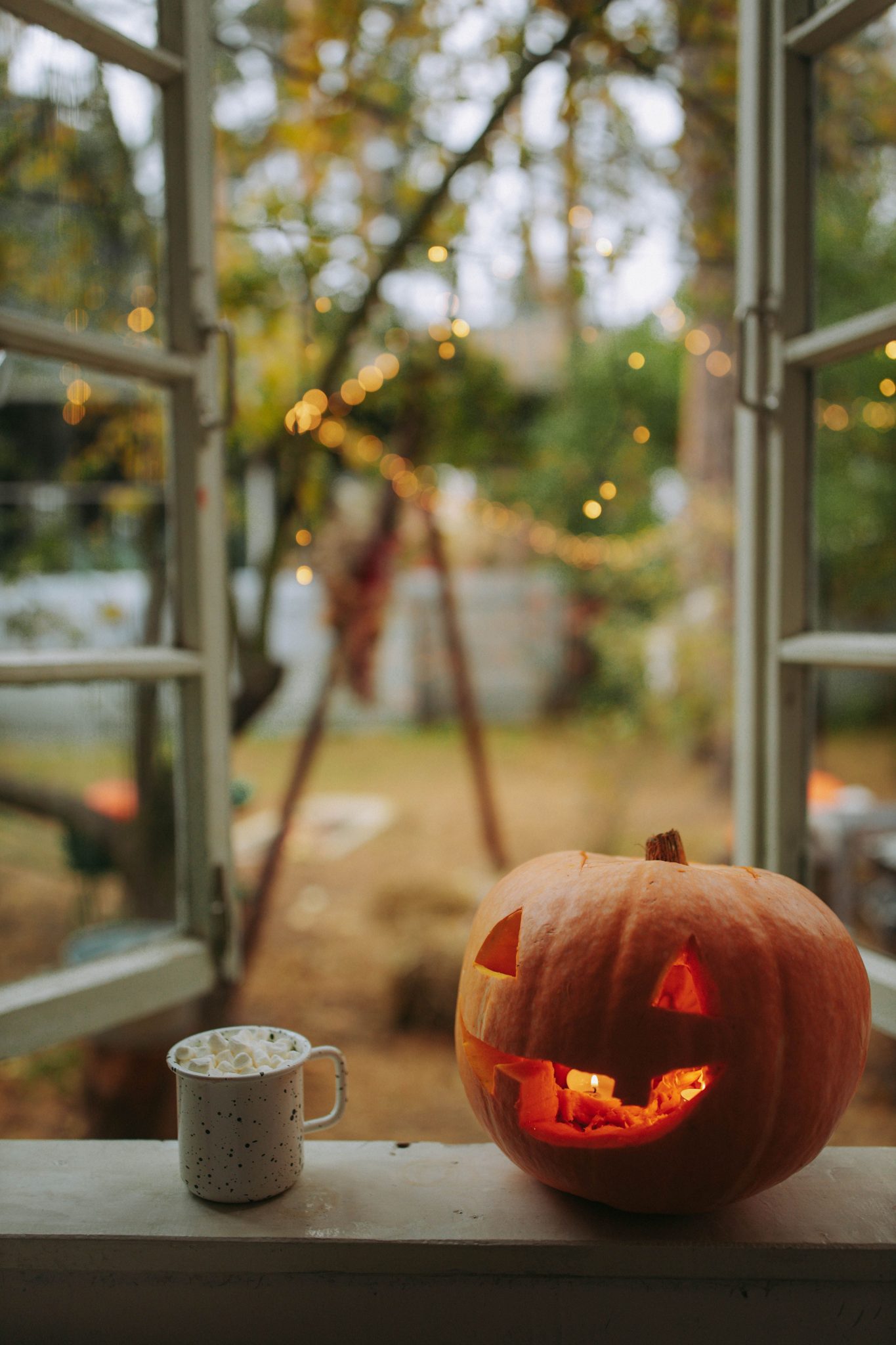 Halloween pumpkin and cozy mug on a window sill with string lights and fall scenery.