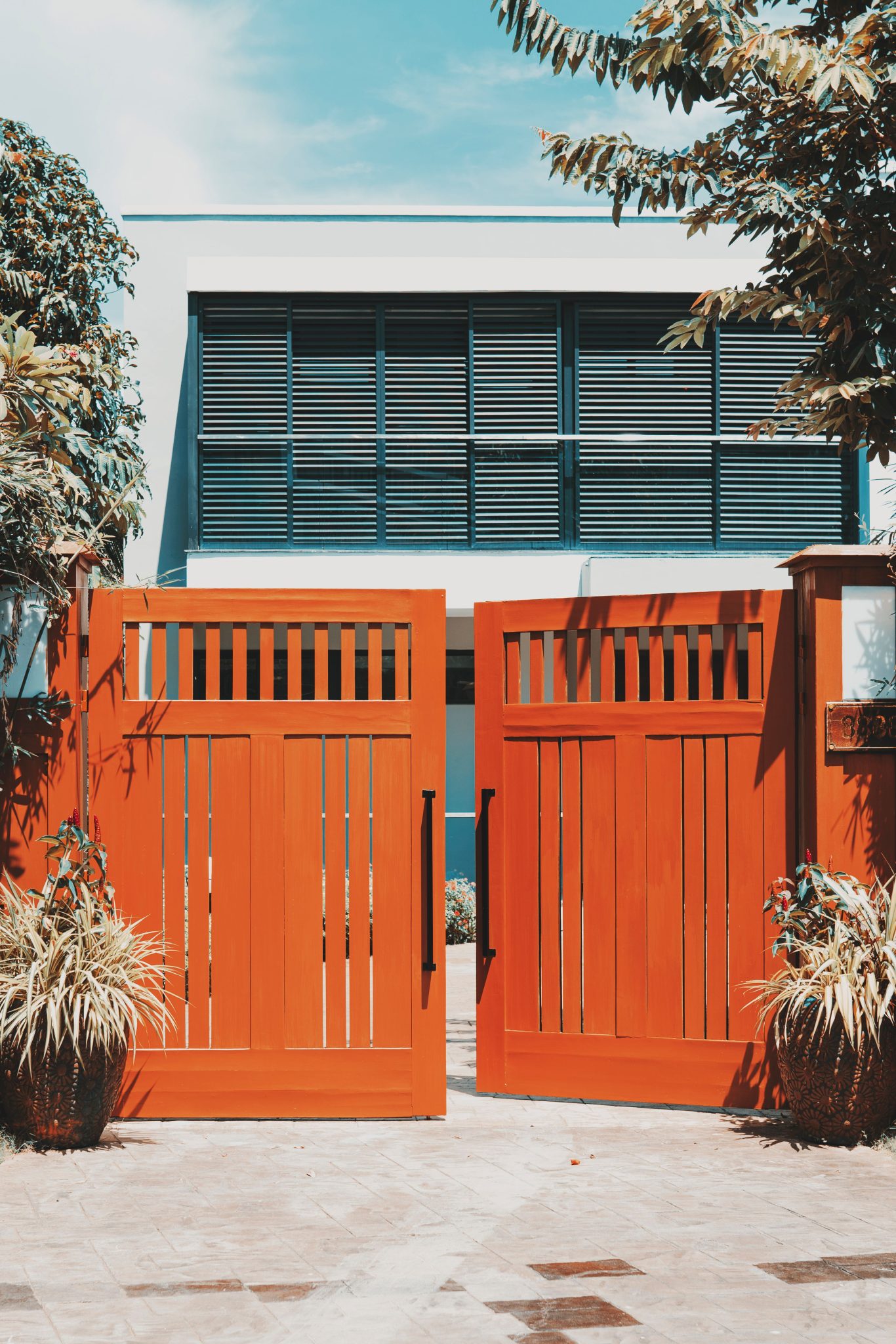Stylish wooden gates open to reveal a modern villa facade, surrounded by lush tropical plants.