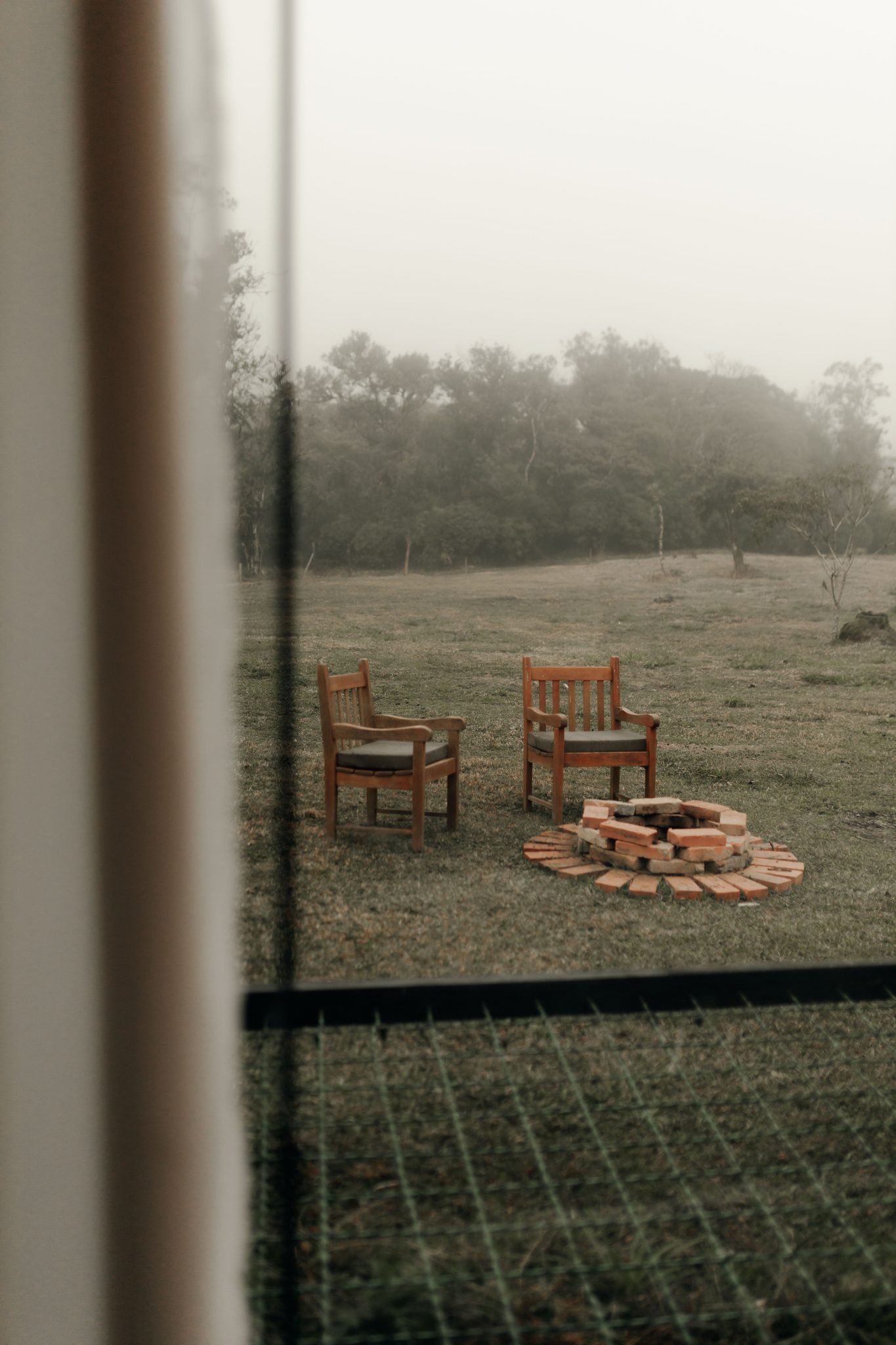 Two wooden chairs encircle a brick fire pit in a foggy outdoor setting, conveying tranquility.