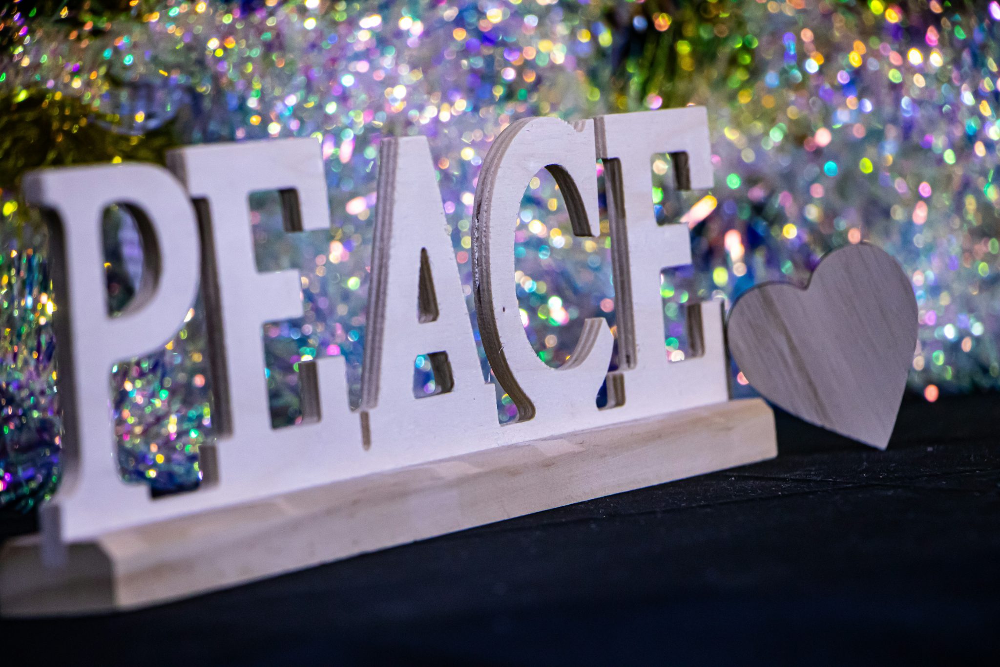 Wooden letters spelling peace and a heart shape stand before a bokeh background of colorful shimmering lights.
