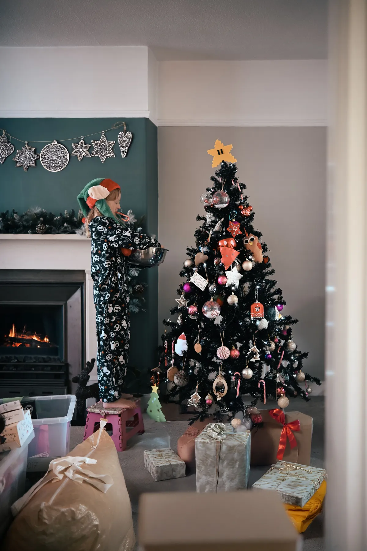 A child dressed as an elf decorates a Christmas tree by a cozy fireplace.