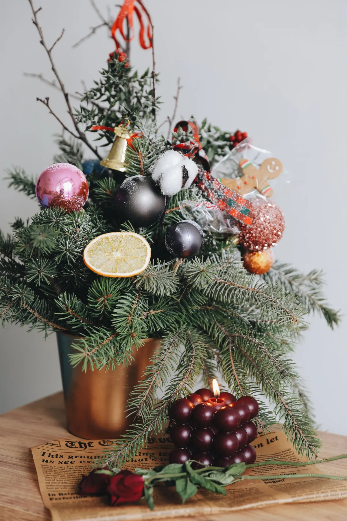 A Christmas floral arrangement with evergreen branches, ornaments, and a burning candle set on a wooden table.