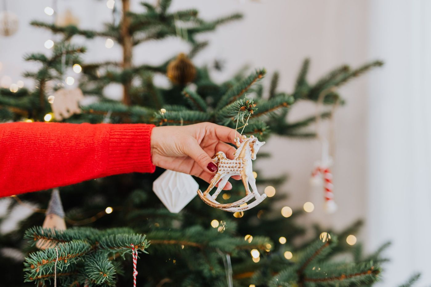 A close-up of a hand hanging a rocking horse ornament on a decorated Christmas tree with twinkling lights.