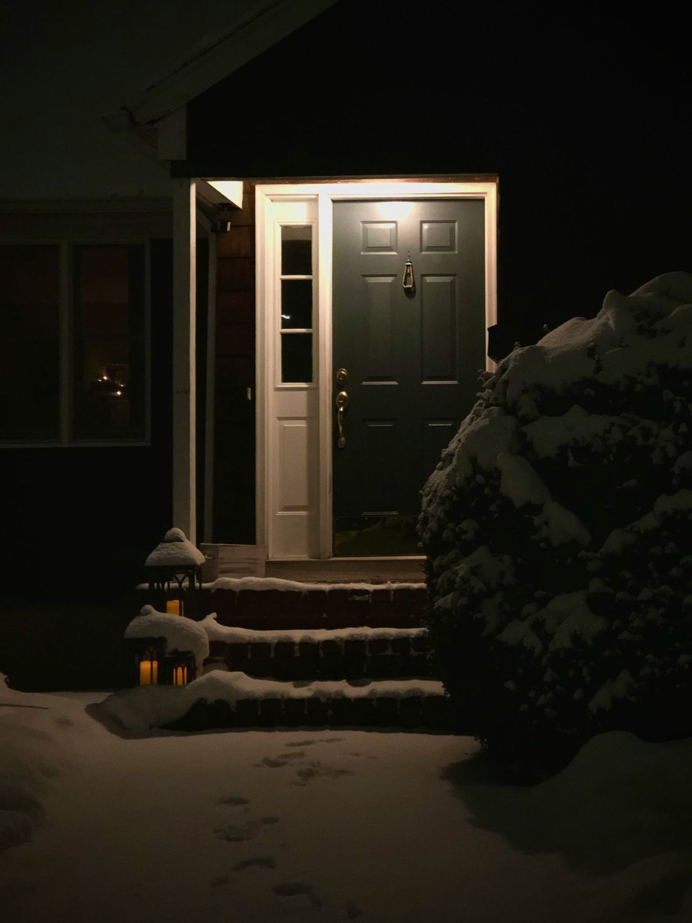 A dark colored front door with an old brass door knocker is illuminated in the darkness of a fresh snowfall.