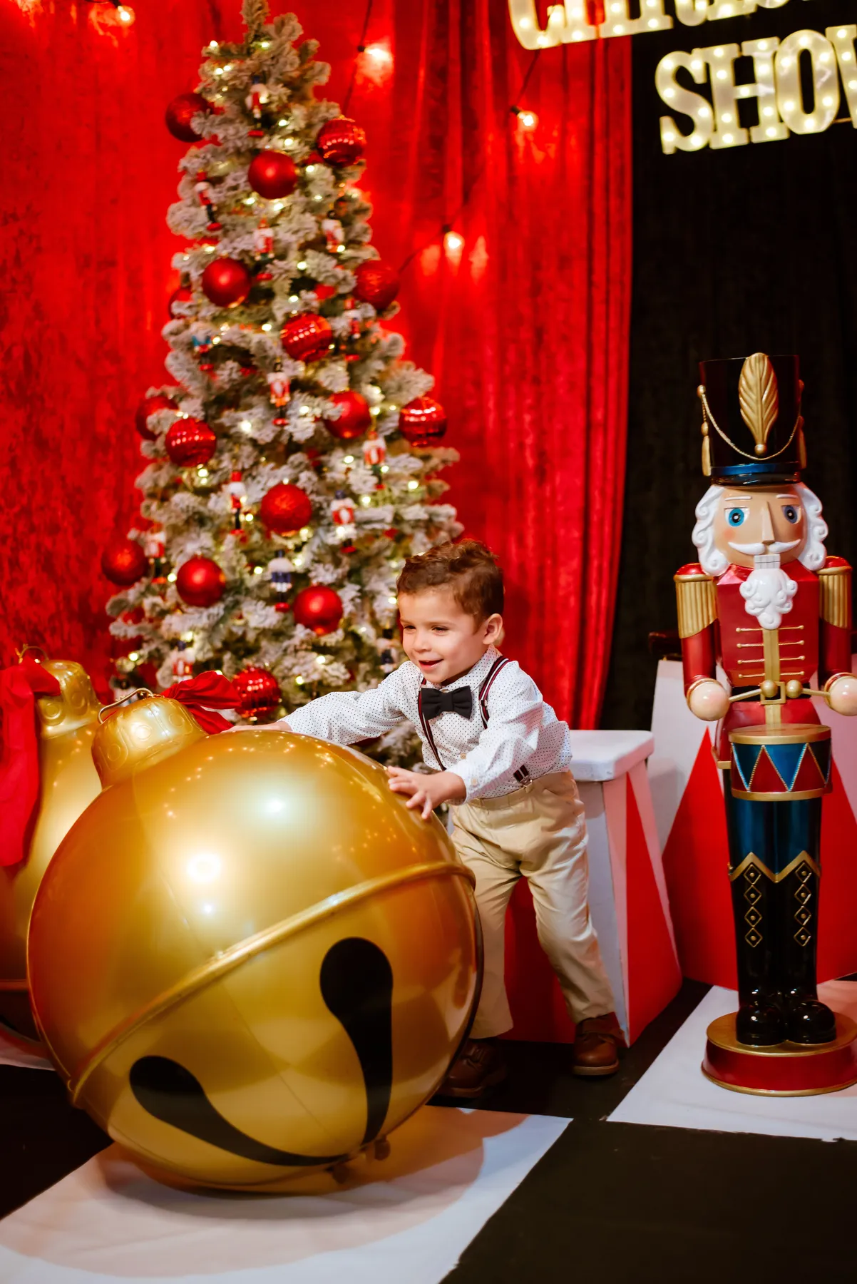 A joyful child playing with a giant golden bell beside a Christmas tree and nutcracker statue.