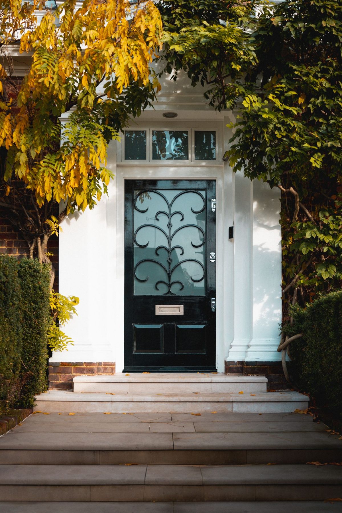 A stylish black door adorned with decorative elements surrounded by autumn foliage in a London neighborhood.