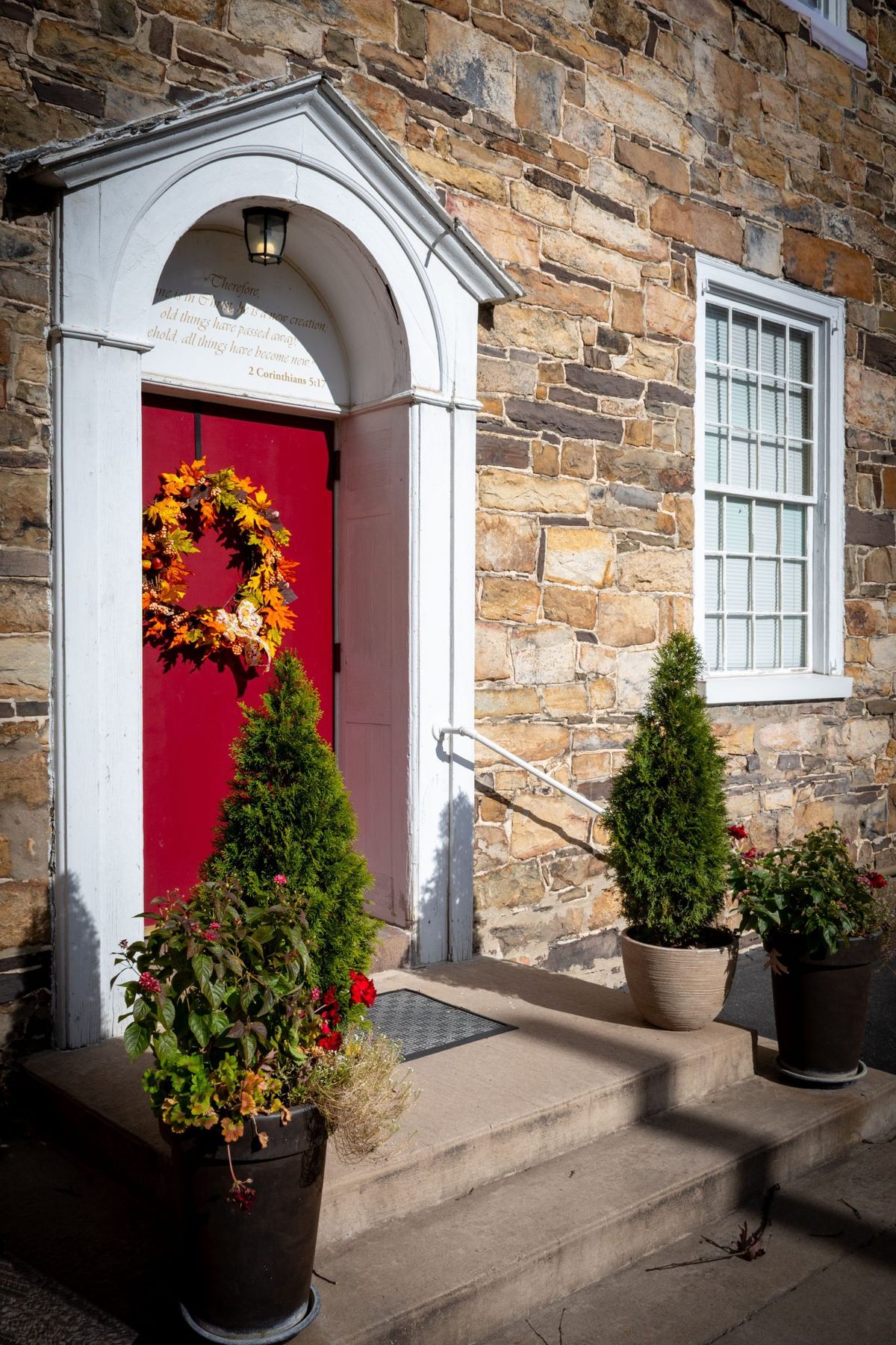 A vibrant red door with fall wreath and potted plants on stone building.