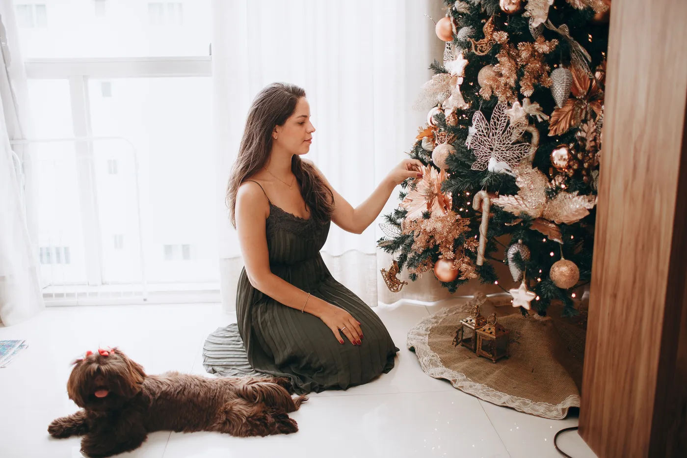 A woman in a green dress decorates a Christmas tree with her dog nearby.