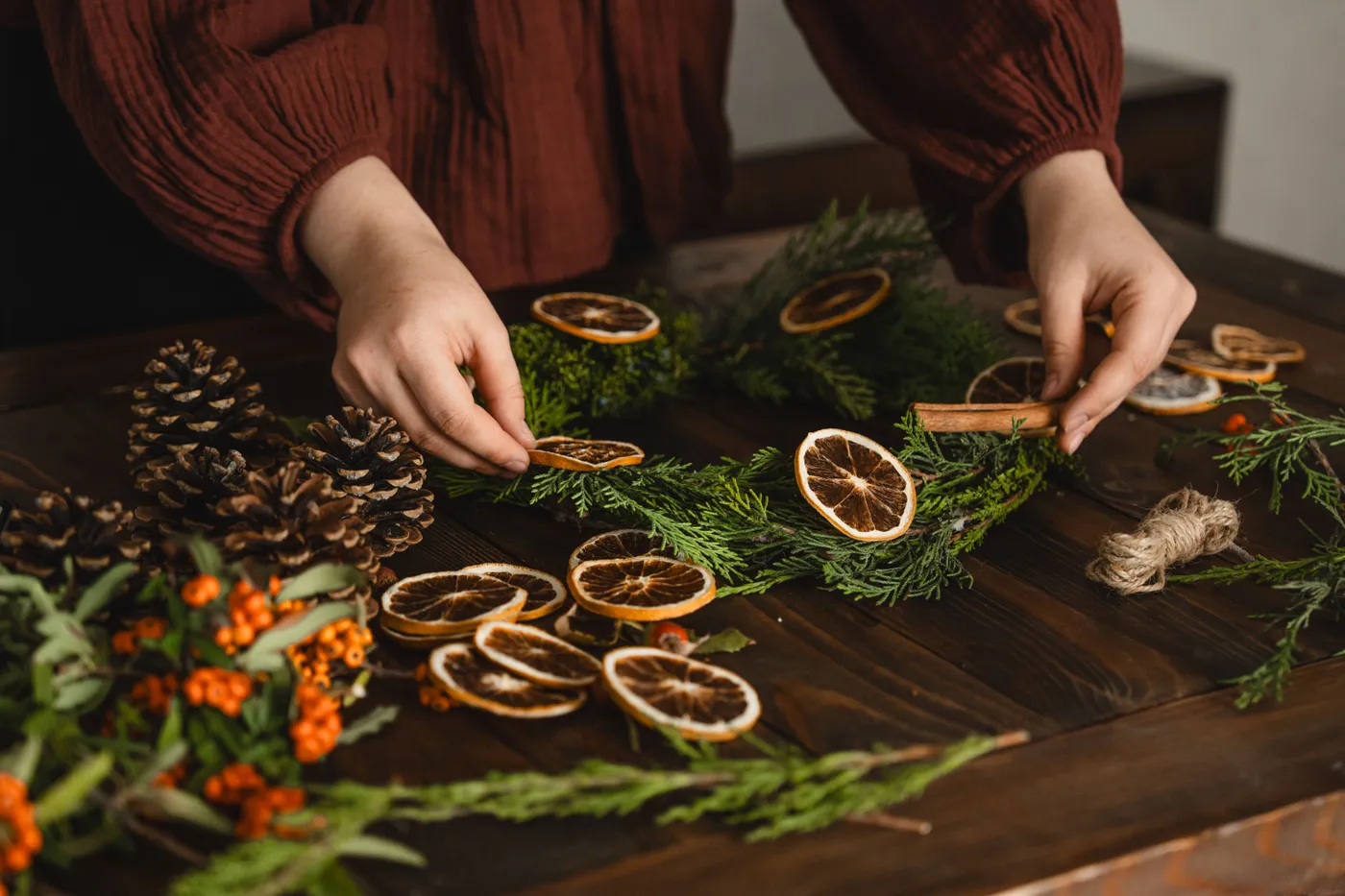 Hands crafting a Christmas wreath with dried oranges, pinecones, and greenery.