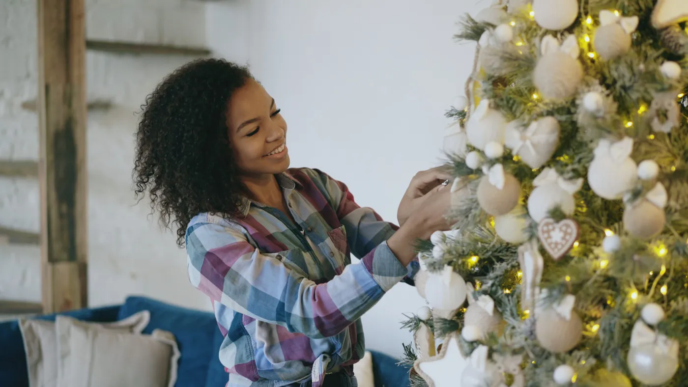 Smiling woman decorates a cozy Christmas tree indoors, adding festive ornaments.