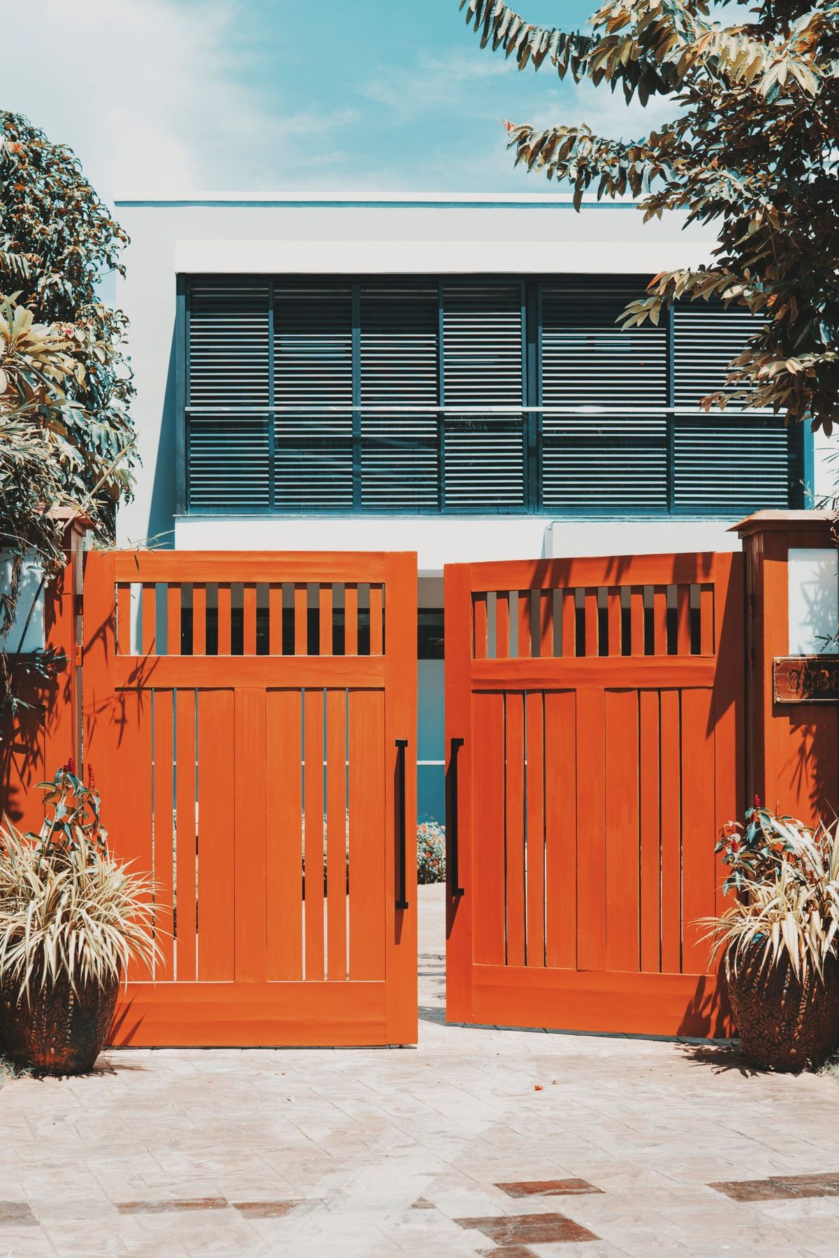 Stylish wooden gates open to reveal a modern villa facade, surrounded by lush tropical plants.