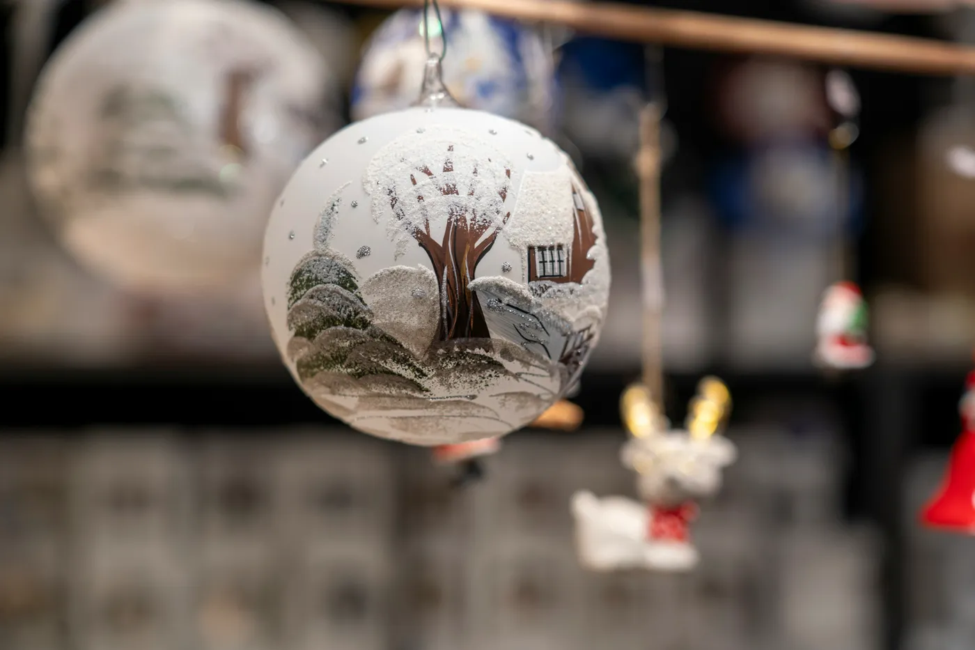 White Christmas Ornament with a Snowy Cottage Design A delicate white Christmas ornament hand-painted with a serene snowy cottage scene, featuring frosted trees and glittering accents. This elegant de