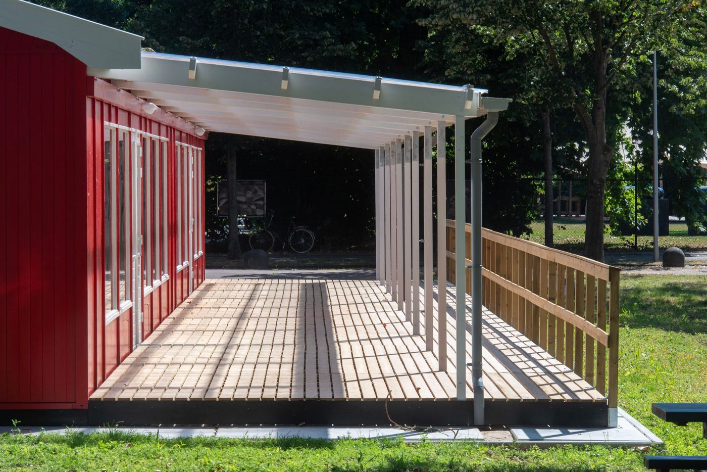 Wooden patio attached to a red building with a transparent roof, outdoor daytime setting.